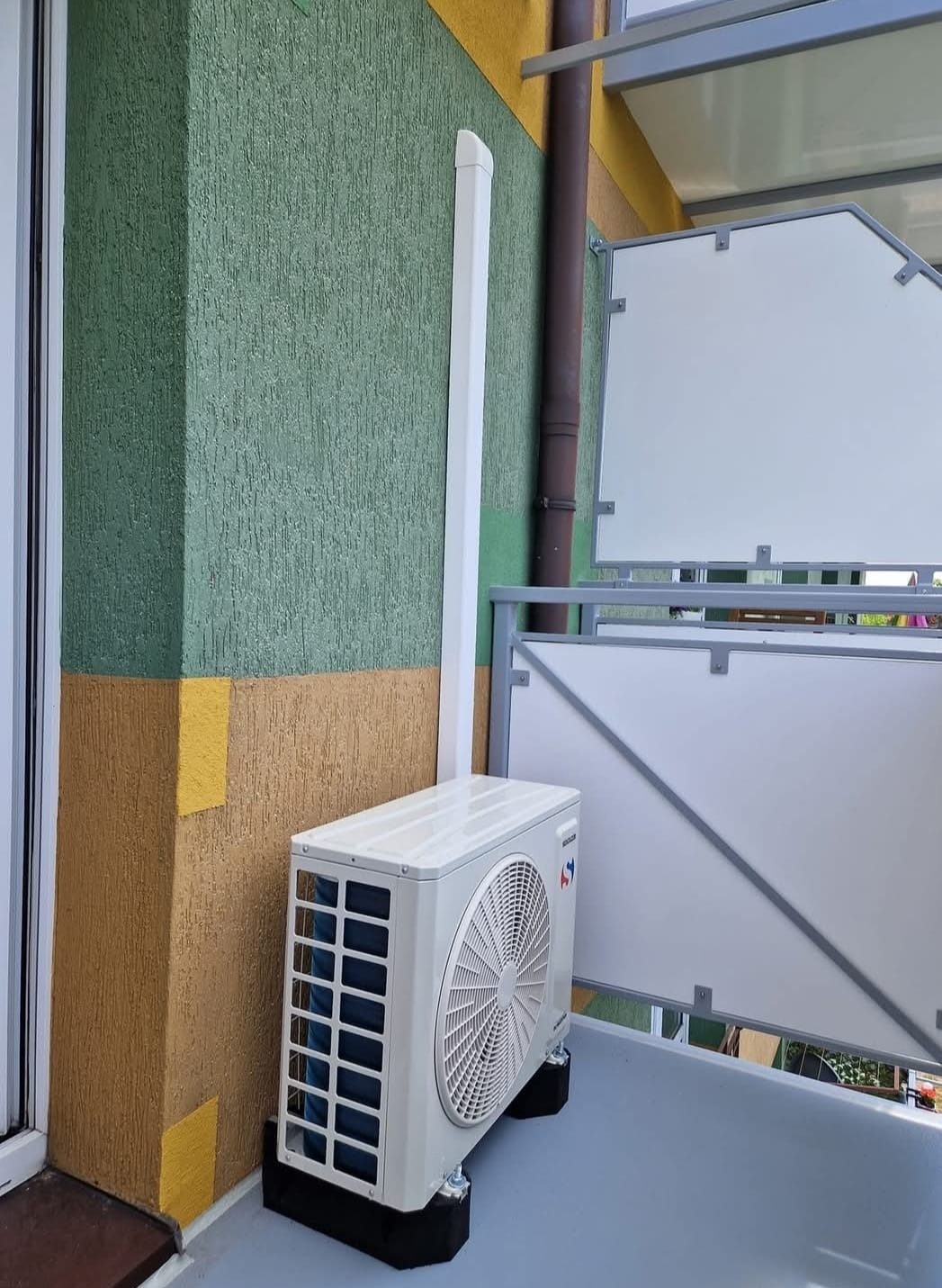 Biała jednostka zewnętrzna klimatyzacji na balkonie, z widoczną rurą odprowadzającą skropliny, na tle ściany w odcieniach zieleni i brązu. Widoczne elementy balustrady.