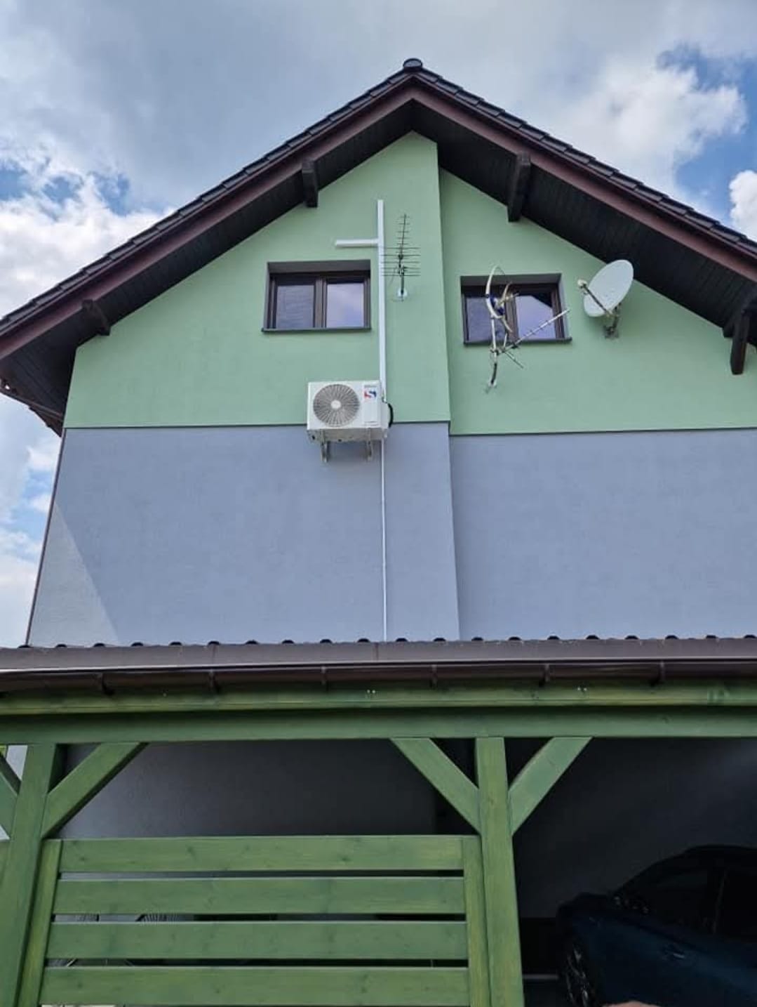 Biały klimatyzator na elewacji dwukolorowego domu z antenami i satelitą, widok z poziomu zadaszenia garażu. Instalacja na ścianie w kolorze szaro-zielonym.