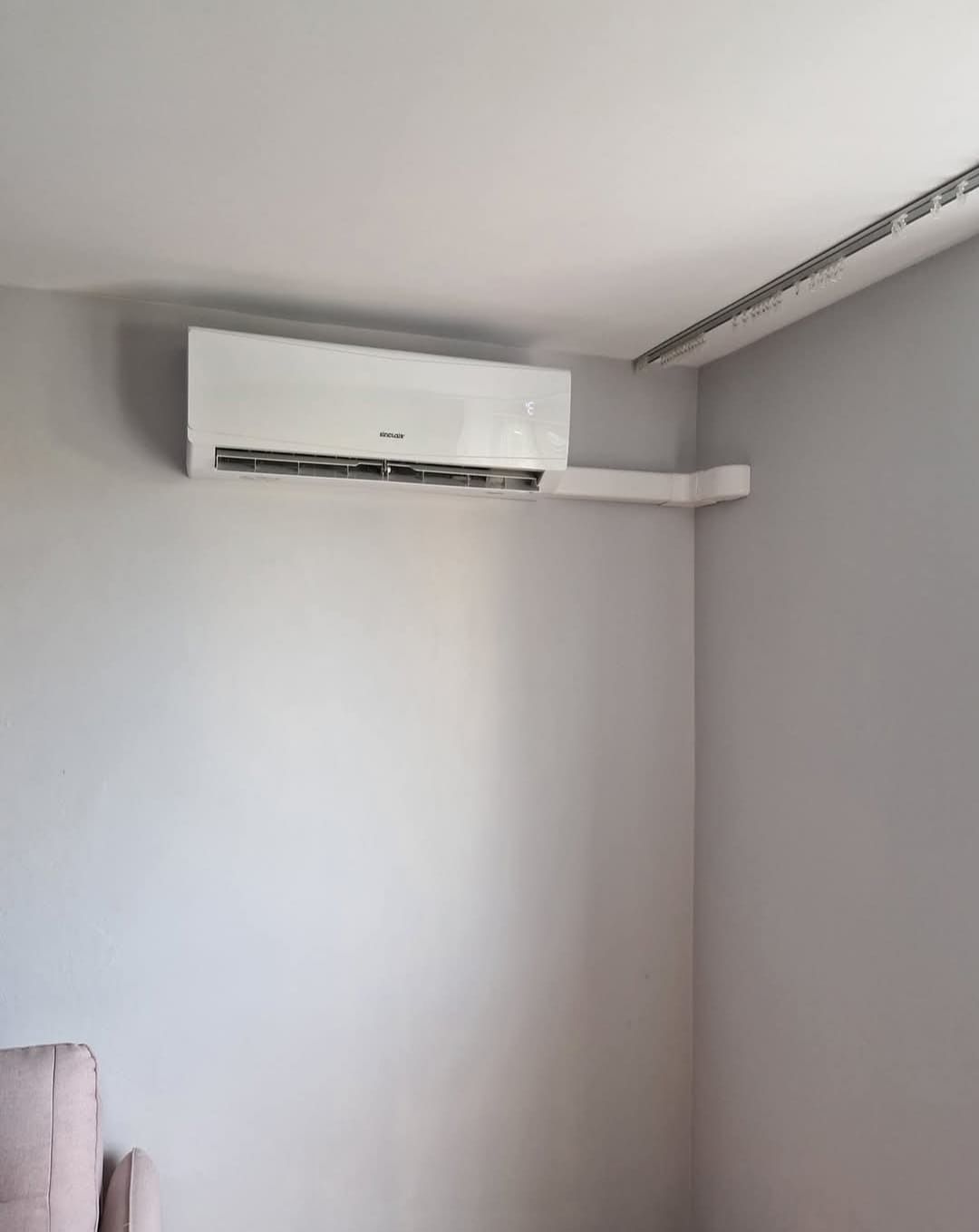 Biały klimatyzator ścienny marki Sinclair zamontowany w rogu szarego pokoju, widoczny fragment szyny oświetleniowej na suficie i fragment sofy w lewym dolnym rogu.