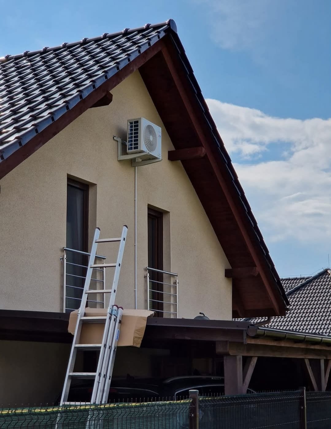 Klimatyzator zamontowany na elewacji domu jednorodzinnego, widoczna drabina oparta o ścianę. Błękitne niebo w tle. Ujęcie z dołu.