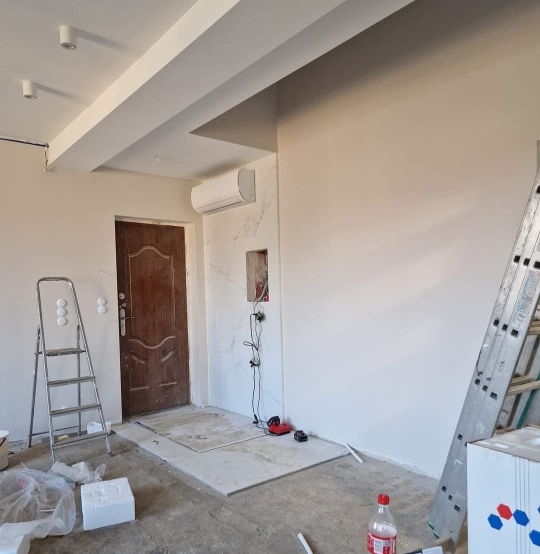 Białe wnętrze w trakcie remontu: zamontowana klimatyzacja, położone płytki podłogowe, ściany przygotowane do malowania, drabina i narzędzia.