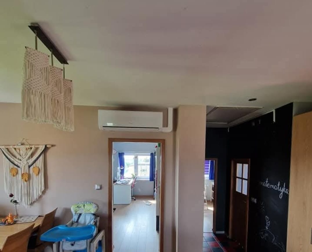 Biała klimatyzacja ścienna w jasnym pokoju z dekoracjami makramowymi i widokiem na inne pomieszczenie. Widoczny fragment sufitu i ściany z tablicą.