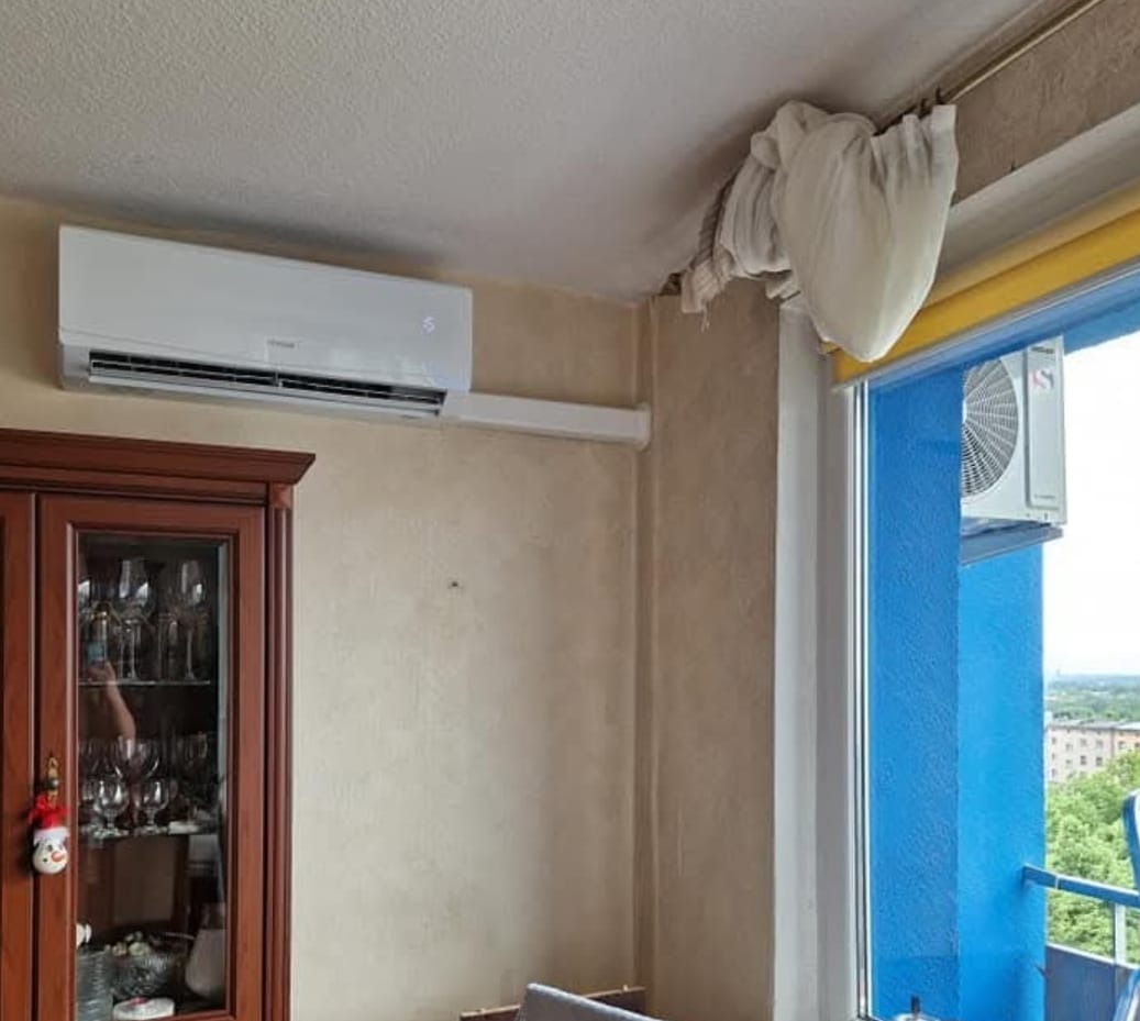 Biały klimatyzator ścienny w pokoju z brązową komodą i niebieskim balkonem. Widoczny drugi klimatyzator na zewnątrz, za oknem.