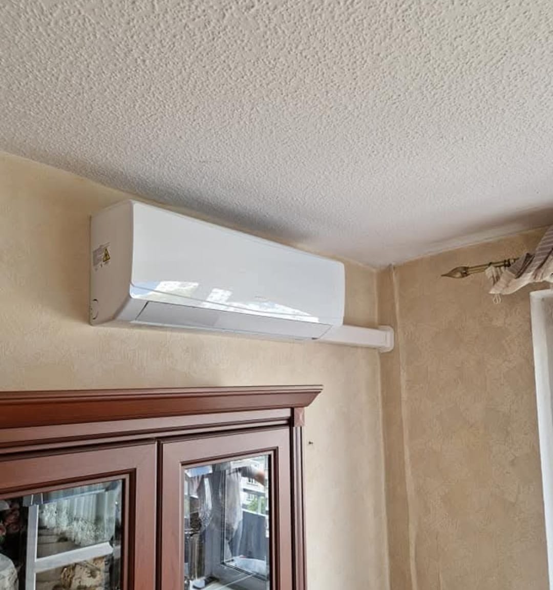 Biała klimatyzacja ścienna w jasnym wnętrzu, widoczna nad ciemnym kredensem. Ściana o fakturze, sufit z widoczną strukturą. Element dekoracyjny okna po prawej.