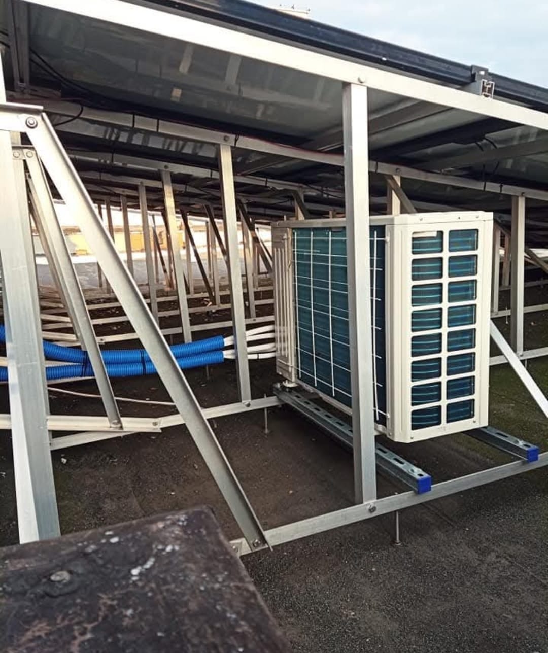 Biała jednostka zewnętrzna klimatyzacji zamontowana na metalowej konstrukcji wsporczej z niebieskimi rurami w tle, widok z dołu.