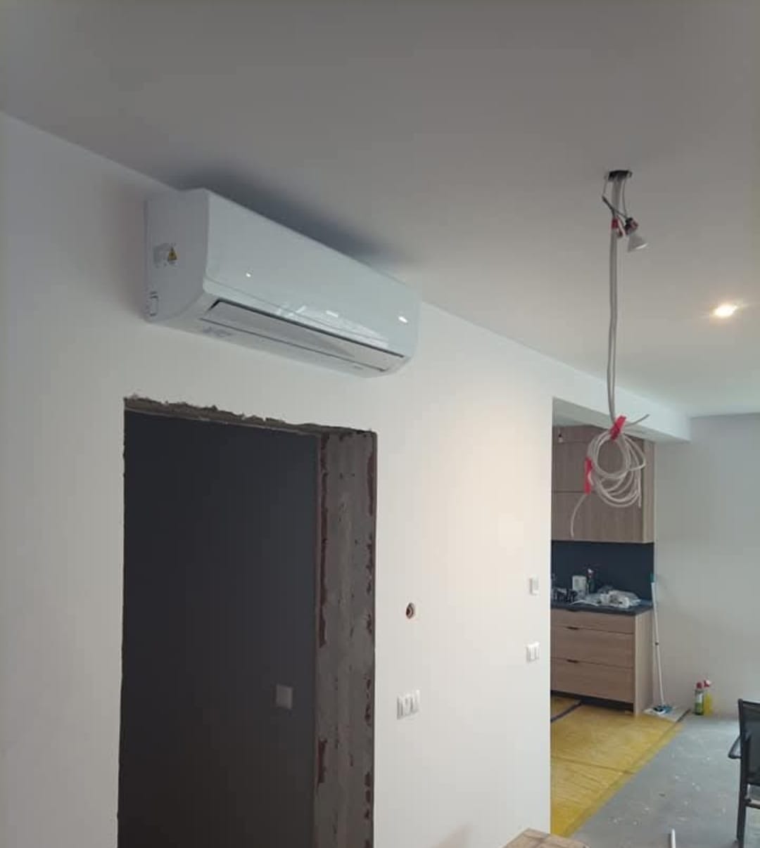 Biały klimatyzator ścienny w jasnym wnętrzu, widoczny otwór drzwiowy w trakcie wykończenia i fragment kuchni w tle. Oświetlenie sufitowe w trakcie montażu.