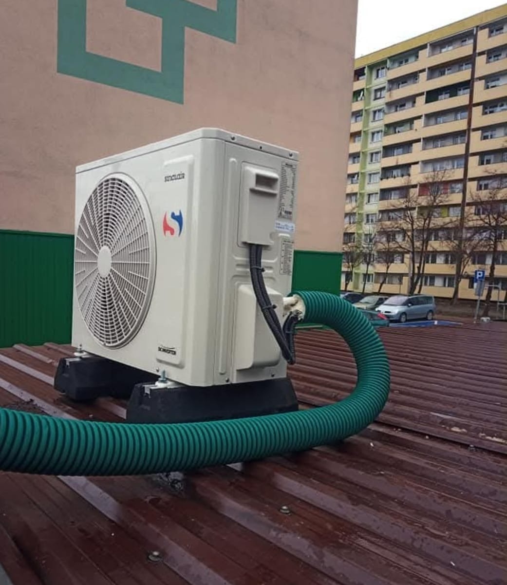 Biała jednostka zewnętrzna klimatyzacji Sinclair na dachu budynku, z widocznymi przewodami i zielonym wężem odprowadzającym skropliny. W tle blok mieszkalny.