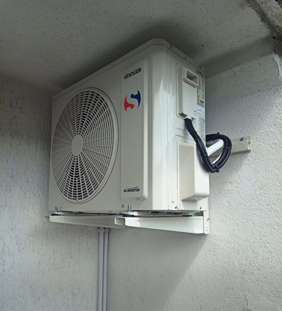 Biała jednostka zewnętrzna klimatyzacji Sinclair zamontowana na szarej ścianie budynku. Widoczne przewody w peszlu i logo producenta.