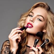 Kobieta z blond włosami, w trakcie makijażu ust czerwoną szminką, trzymająca pędzle do makijażu. Delikatny makijaż oczu, koronkowy top.