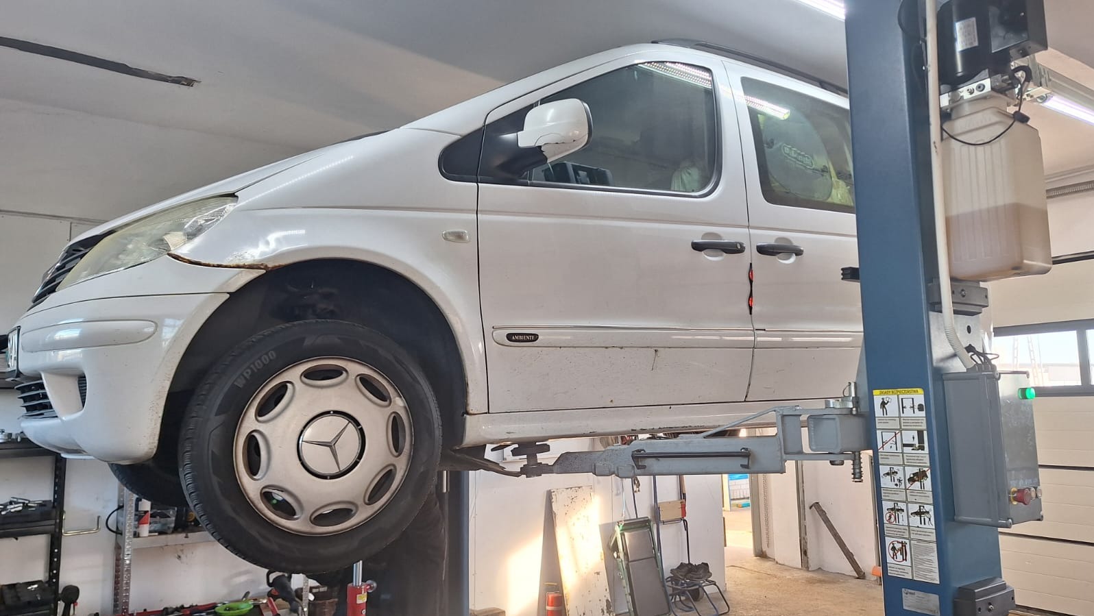 Biały Mercedes Ambiente na podnośniku w warsztacie. Widoczne ślady korozji na karoserii, oświetlenie warsztatowe i elementy wyposażenia. Ujęcie z boku, z bliska.
