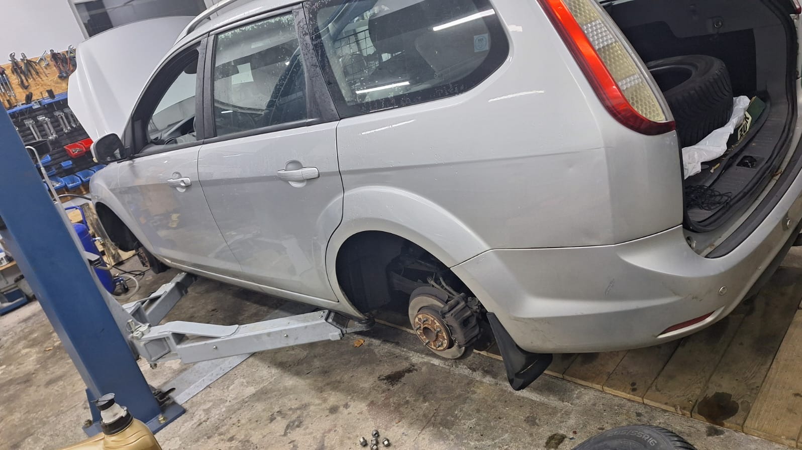 Srebrny samochód kombi na podnośniku w warsztacie, z otwartą maską i zdjętym kołem, widoczne narzędzia i opona w bagażniku. Przegląd lub naprawa zawieszenia.