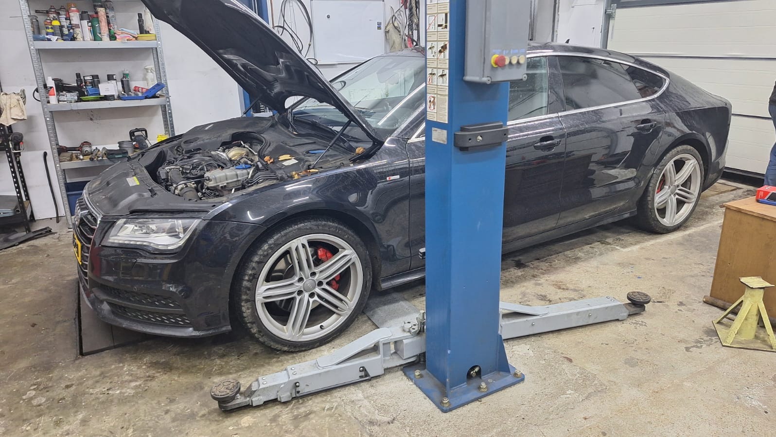 Czarny samochód Audi A7 na podnośniku w warsztacie z otwartą maską, widoczne elementy silnika i narzędzia na półce w tle. Czerwone zaciski hamulcowe.