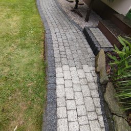 CLEAN WALL NON PROFIT SPÓŁKA Z OGRANICZONĄ ODPOWIEDZIALNOŚCIĄ - Zakrzywiony chodnik z jasnoszarej kostki brukowej, obrzeża z ciemnoszarej kostki, trawnik po lewej, rośliny i schody po prawej stronie.