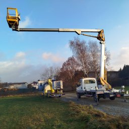 CLEAN WALL NON PROFIT SPÓŁKA Z OGRANICZONĄ ODPOWIEDZIALNOŚCIĄ - Dwa podnośniki koszowe na trawiastym terenie, jeden z wysięgnikiem uniesionym ku niebu, drugi zaparkowany obok, w tle zabudowania mieszkalne i błękitne niebo.