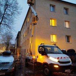 CLEAN WALL NON PROFIT SPÓŁKA Z OGRANICZONĄ ODPOWIEDZIALNOŚCIĄ - Żółty podnośnik koszowy na białym samochodzie Iveco, oświetlony, na tle budynku z zapalonymi oknami, wczesny wieczór.