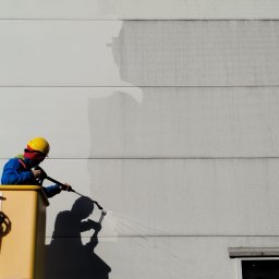 CLEAN WALL NON PROFIT SPÓŁKA Z OGRANICZONĄ ODPOWIEDZIALNOŚCIĄ - Pracownik w żółtym kasku myje elewację budynku z podnośnika koszowego. Widoczny efekt czyszczenia - różnica między umytą a brudną powierzchnią.