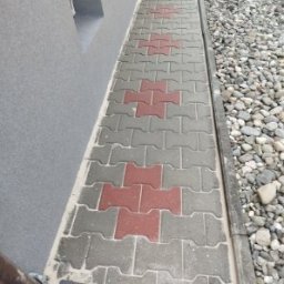 CLEAN WALL NON PROFIT SPÓŁKA Z OGRANICZONĄ ODPOWIEDZIALNOŚCIĄ - Ułożona kostka brukowa wzdłuż ściany budynku, z elementami w kolorze szarym i czerwonym, oddzielona od kamieni ozdobnych wąskim korytkiem.