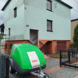 CLEAN WALL NON PROFIT SPÓŁKA Z OGRANICZONĄ ODPOWIEDZIALNOŚCIĄ - Pracownik na rusztowaniu czyści elewację domu z numerem 107 za pomocą myjki ciśnieniowej Dibo. Widoczny efekt czyszczenia - górna część elewacji jest czystsza.