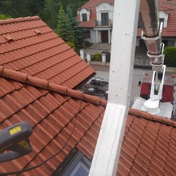 CLEAN WALL NON PROFIT SPÓŁKA Z OGRANICZONĄ ODPOWIEDZIALNOŚCIĄ - Czerwona dachówka widziana z góry, z fragmentem podnośnika koszowego i narzędziem do czyszczenia. W tle dom z ogrodem i drzewami.