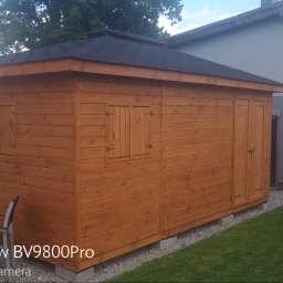 CLEAN WALL NON PROFIT SPÓŁKA Z OGRANICZONĄ ODPOWIEDZIALNOŚCIĄ - Drewniany domek narzędziowy z czarnym dachem, umieszczony na betonowych bloczkach, na tle trawnika i budynku mieszkalnego. Widoczne okiennice i drzwi.