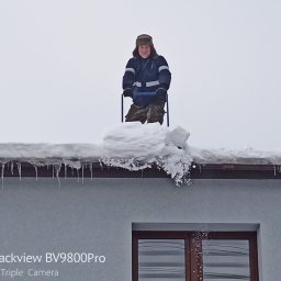 CLEAN WALL NON PROFIT SPÓŁKA Z OGRANICZONĄ ODPOWIEDZIALNOŚCIĄ - Mężczyzna w czapce usuwa śnieg z dachu domu za pomocą specjalnego narzędzia. Widoczne sople lodu i okno. Pochmurna pogoda.