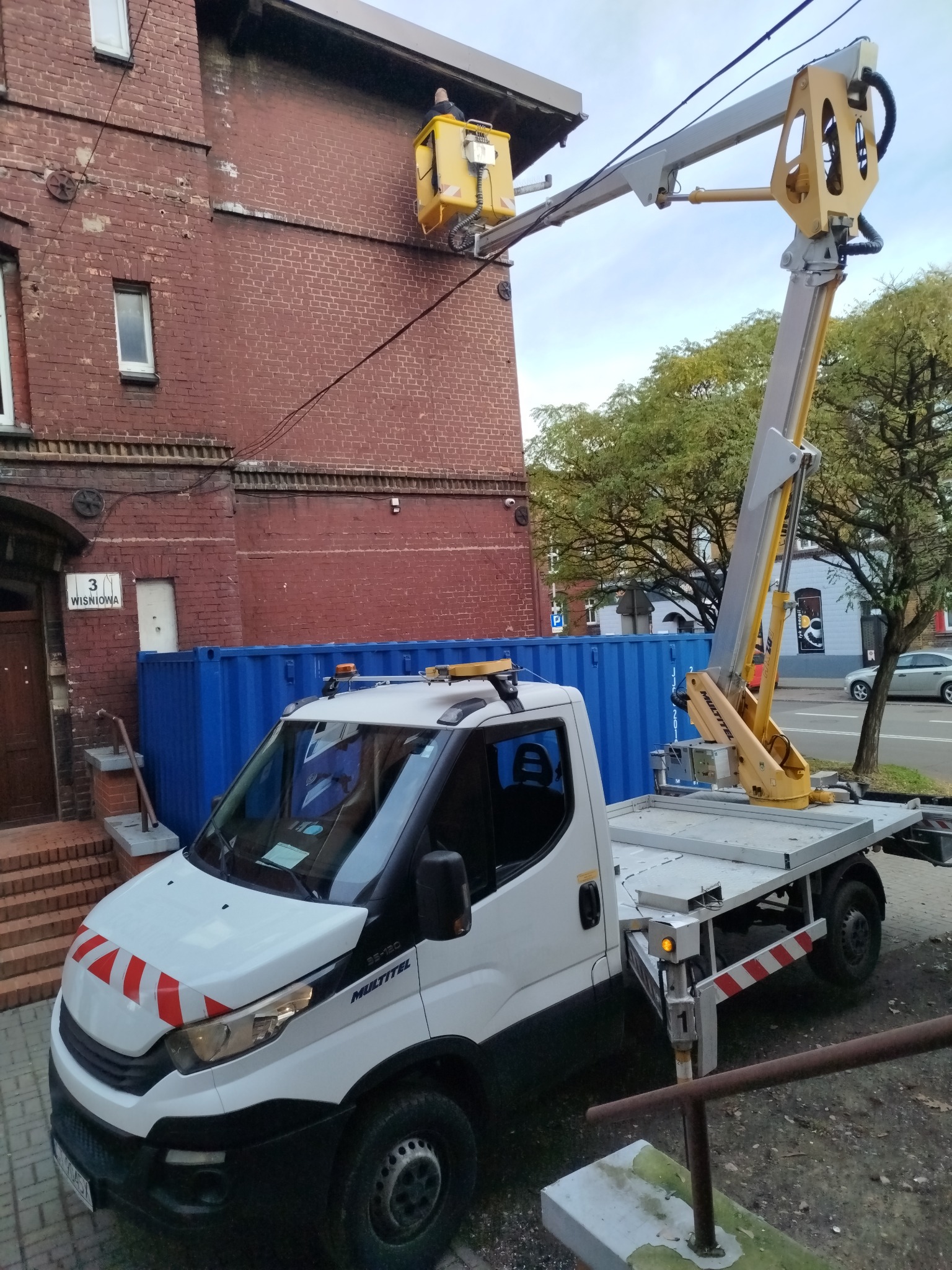 Żółty podnośnik koszowy na tle ceglanej ściany budynku w Sosnowcu. Widoczny fragment ulicy i drzewa. Pojazd z wysięgnikiem zaparkowany przy chodniku.