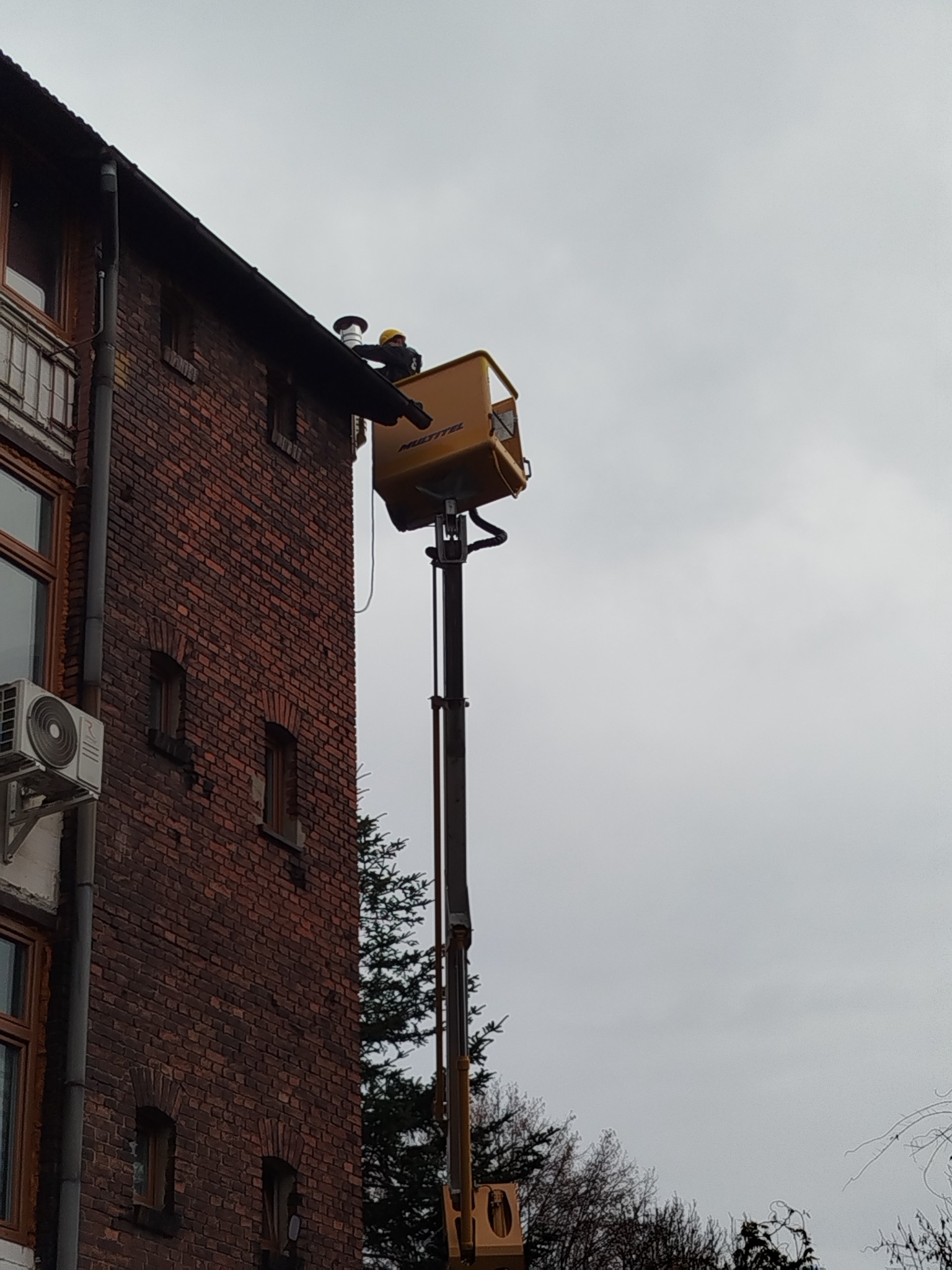 Pracownik w żółtym podnośniku koszowym przy ceglanej ścianie budynku, prawdopodobnie wykonuje prace na wysokości, widoczny fragment dachu i rynny.