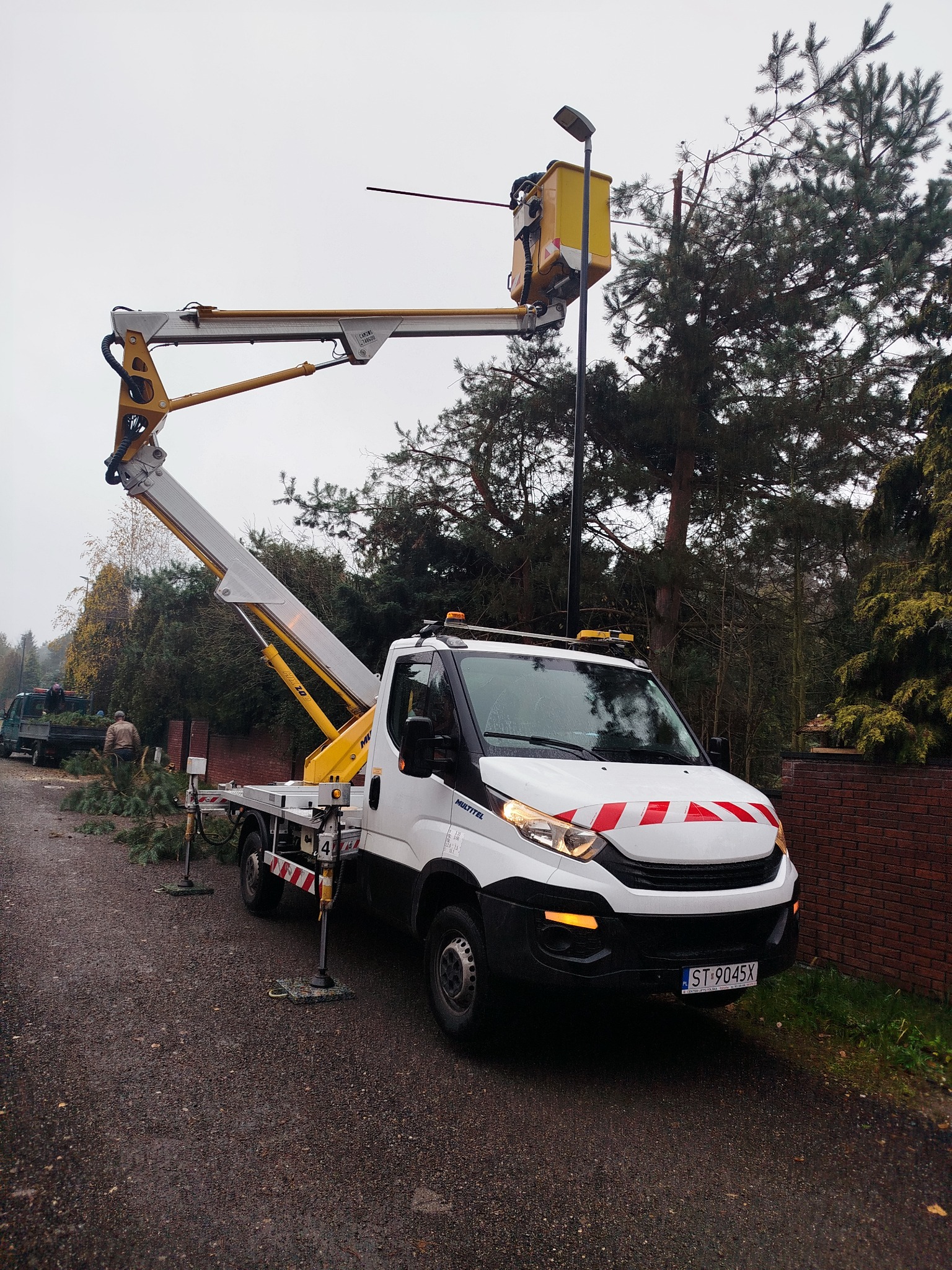 Biały podnośnik koszowy na drodze, z żółtym koszem i wysięgnikiem, obok ściętych gałęzi i drzew. W tle widoczny fragment lasu i ceglany mur.
