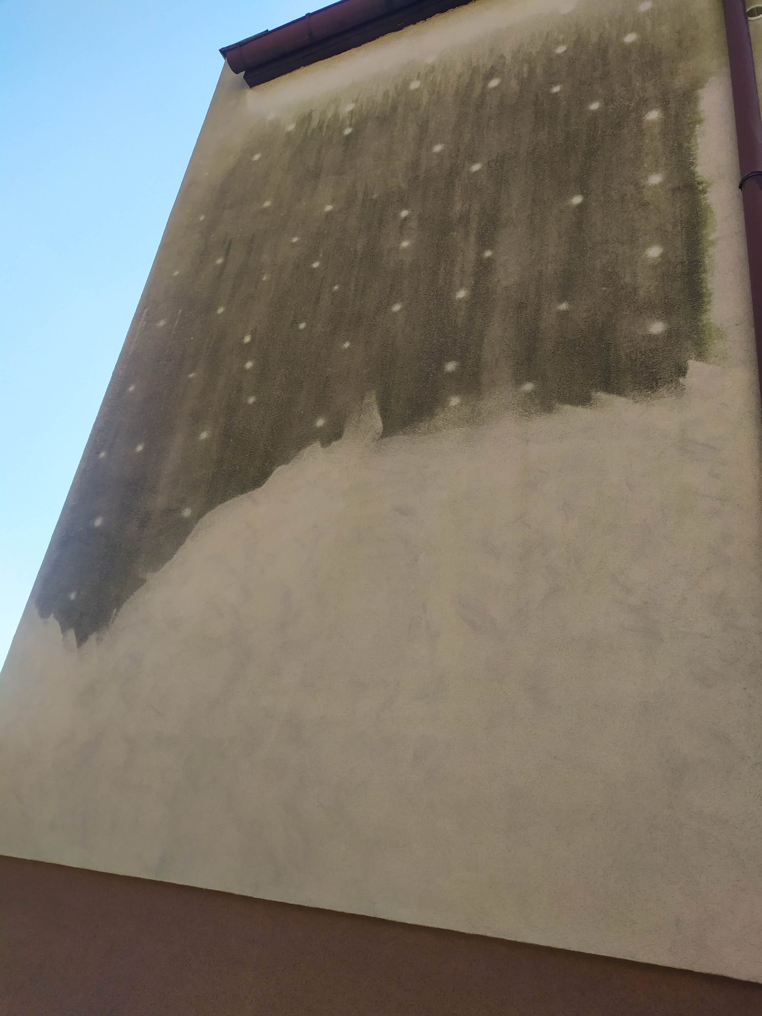 Częściowo oczyszczona elewacja budynku z widocznymi śladami zabrudzeń i punktowymi pozostałościami po czyszczeniu. Ujęcie z dołu, fragment rynny po prawej.