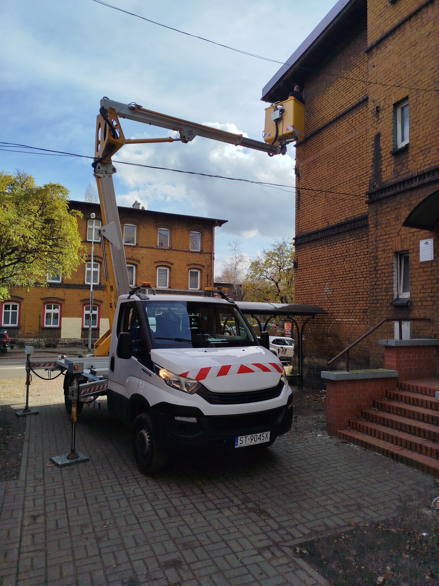 Żółty podnośnik koszowy na chodniku, z pracownikiem w koszu przy ceglanej elewacji budynku. Widoczne stabilizatory i tablica rejestracyjna pojazdu.
