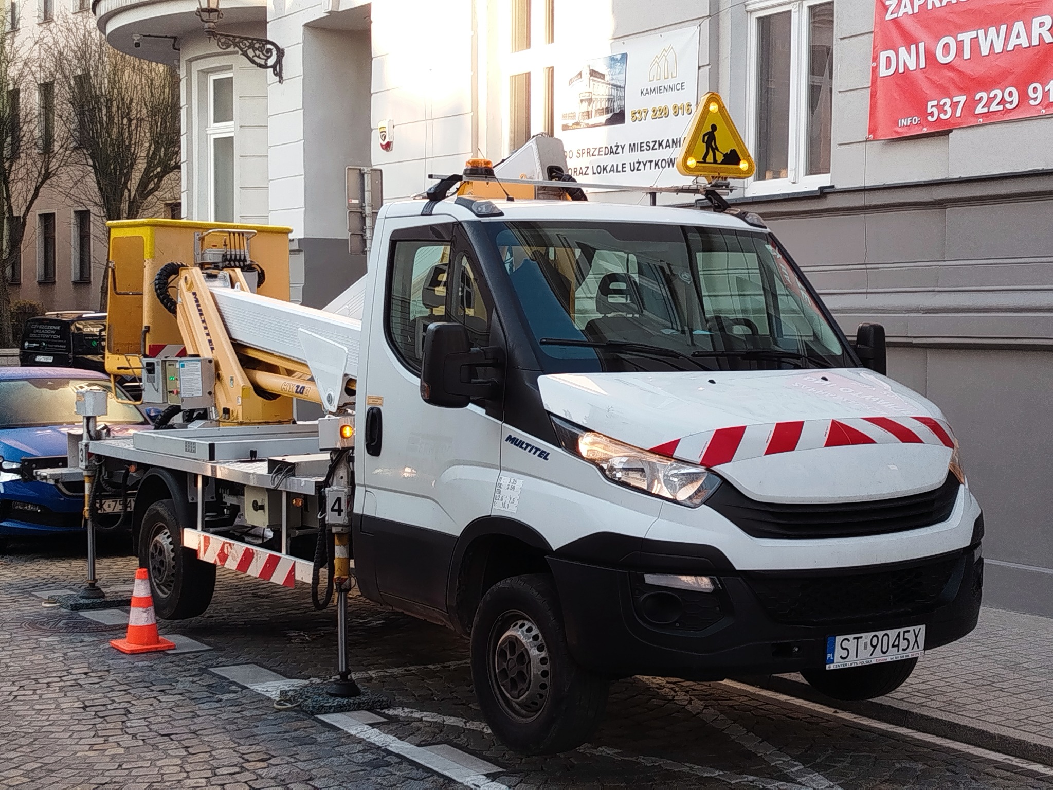 Podnośnik koszowy Multitel na chodniku z kostki, zabezpieczony pachołkiem. Na dachu pojazdu znak ostrzegawczy. W tle budynek z banerem reklamowym.
