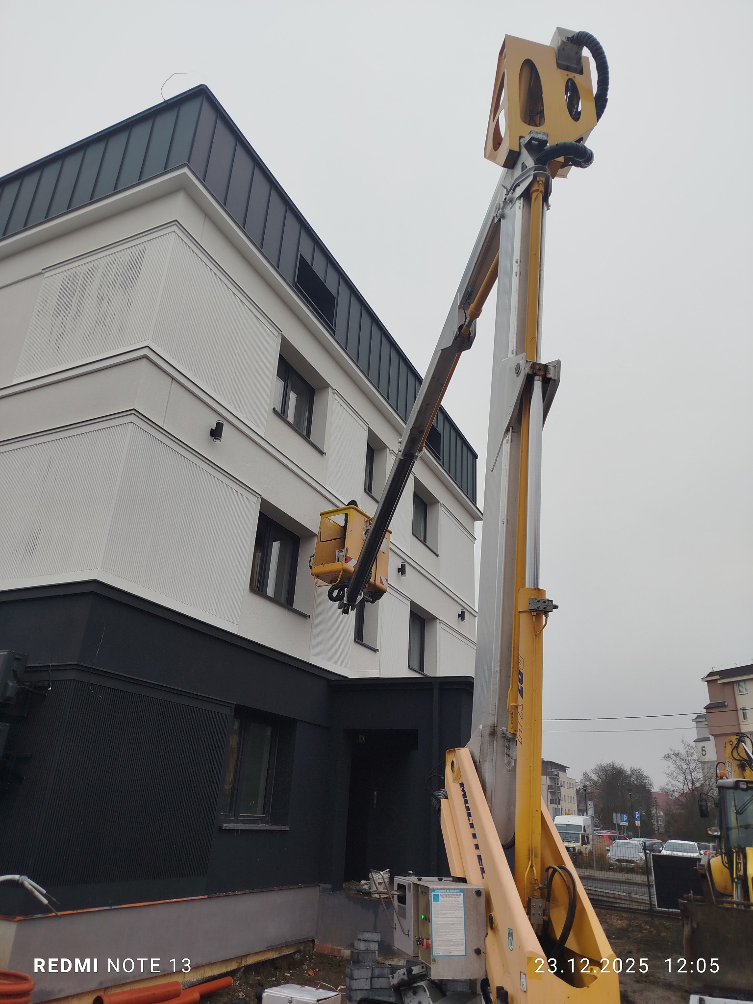 Żółty podnośnik koszowy na tle budynku z białą elewacją i czarnym cokołem. Kosz podnośnika ustawiony przy oknie. Pochmurne niebo w tle.