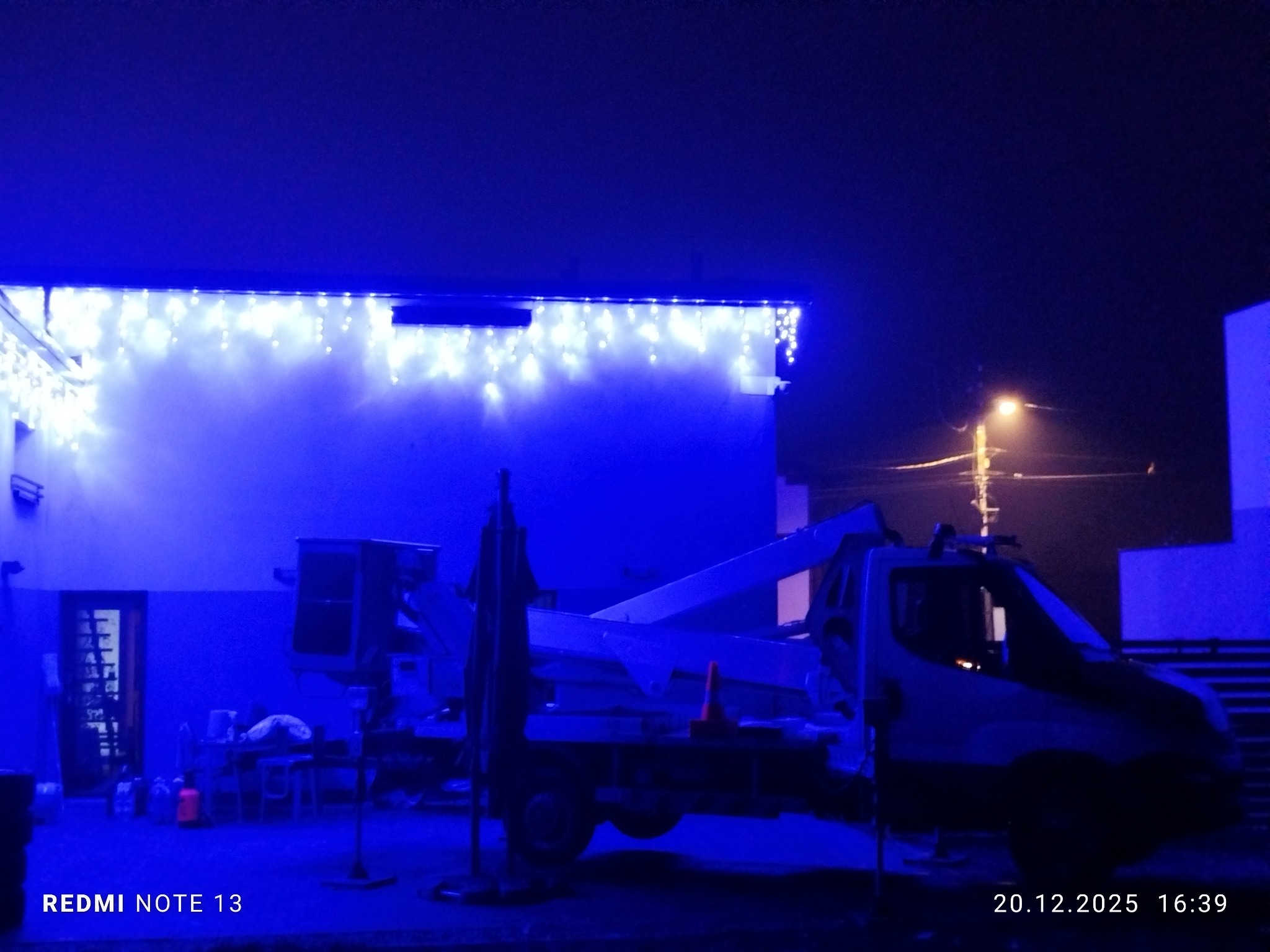 Podnośnik koszowy oświetlony niebieskim światłem, zaparkowany przed budynkiem z dekoracyjnym oświetleniem. Widoczny fragment ulicznej latarni w tle.