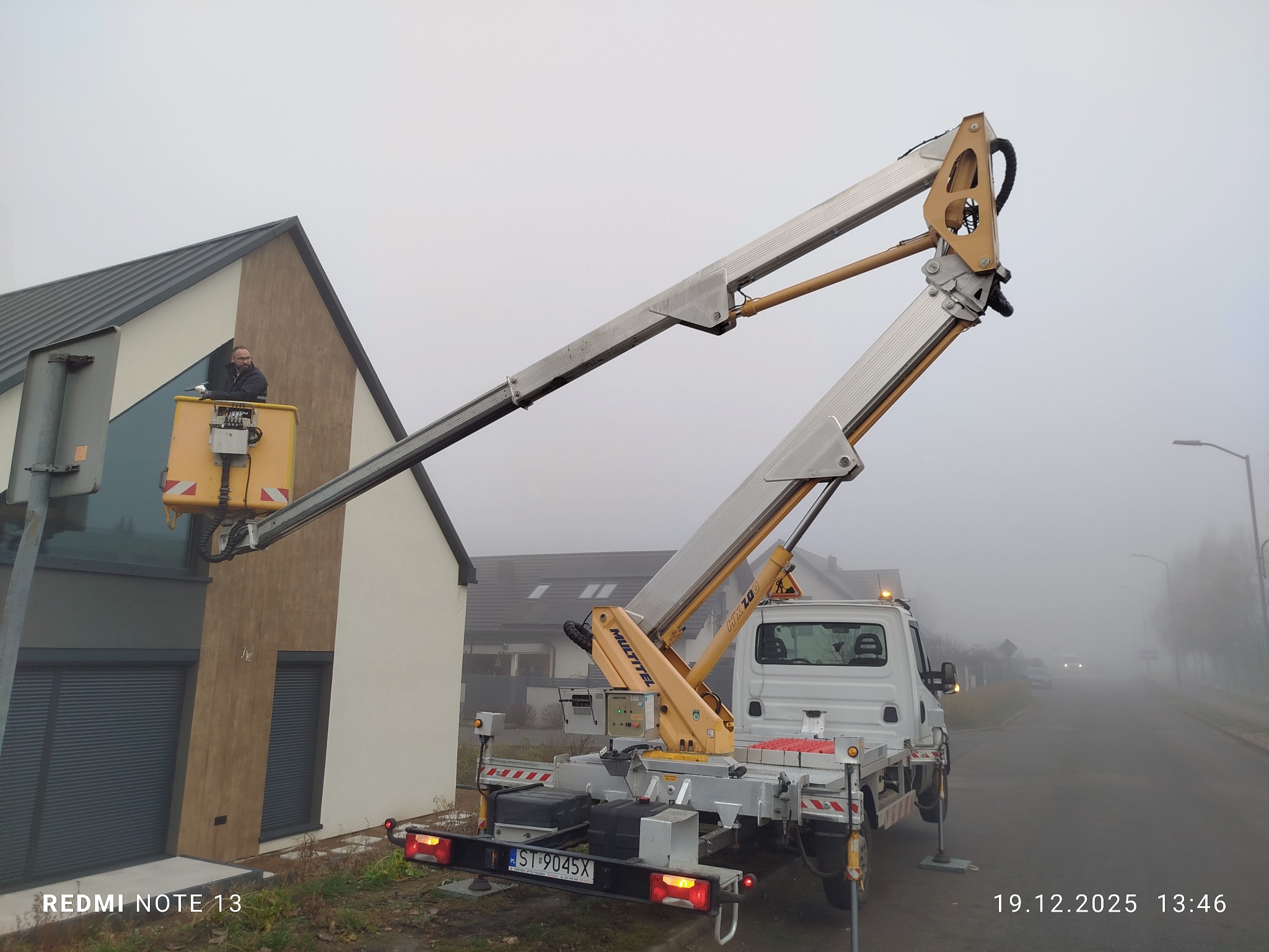 Pracownik w koszu podnośnika koszowego, na tle nowoczesnego domu z elementami drewna, wykonuje prace na wysokości. Mglista pogoda.