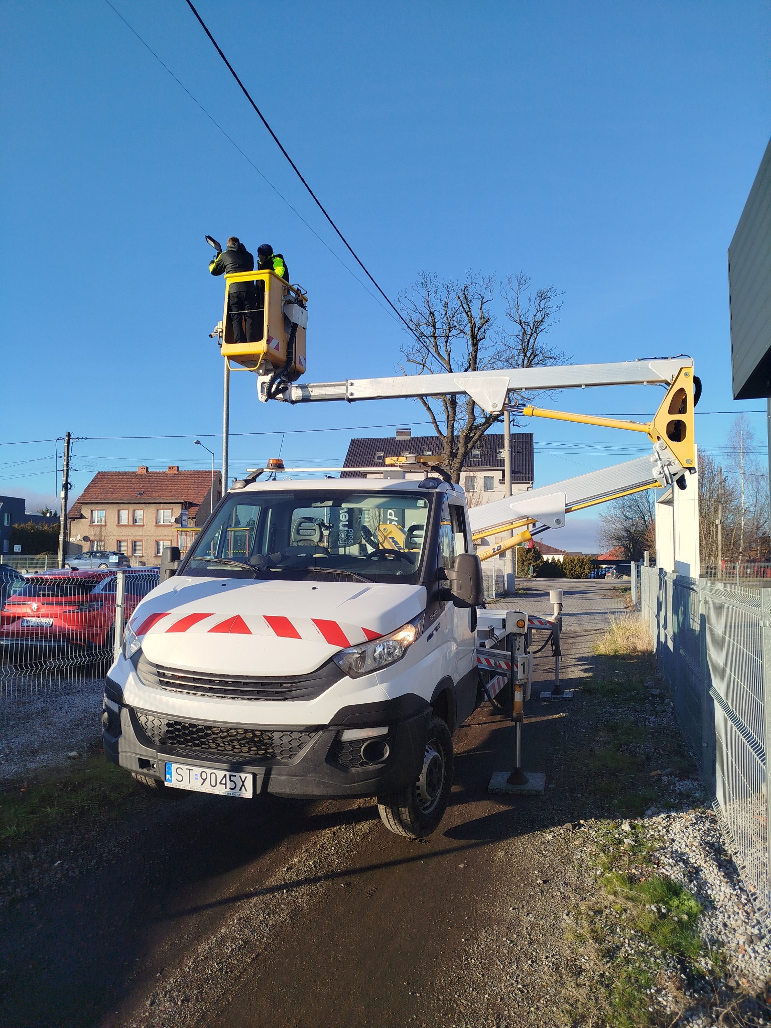 Biały samochód z podnośnikiem koszowym na tle błękitnego nieba, dwóch pracowników w koszu wykonuje prace na wysokości, widoczne budynki w tle.