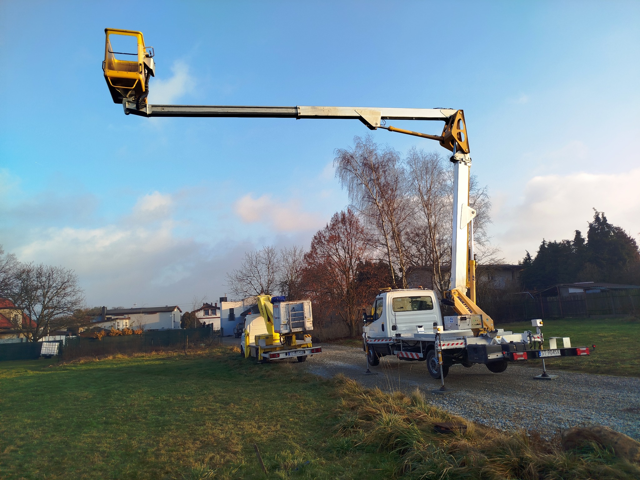 Dwa podnośniki koszowe na trawiastym terenie, jeden z wysięgnikiem uniesionym ku niebu, drugi zaparkowany obok, w tle zabudowania mieszkalne i błękitne niebo.