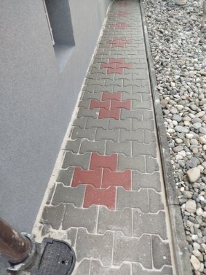 Ułożona kostka brukowa wzdłuż ściany budynku, z elementami w kolorze szarym i czerwonym, oddzielona od kamieni ozdobnych wąskim korytkiem.
