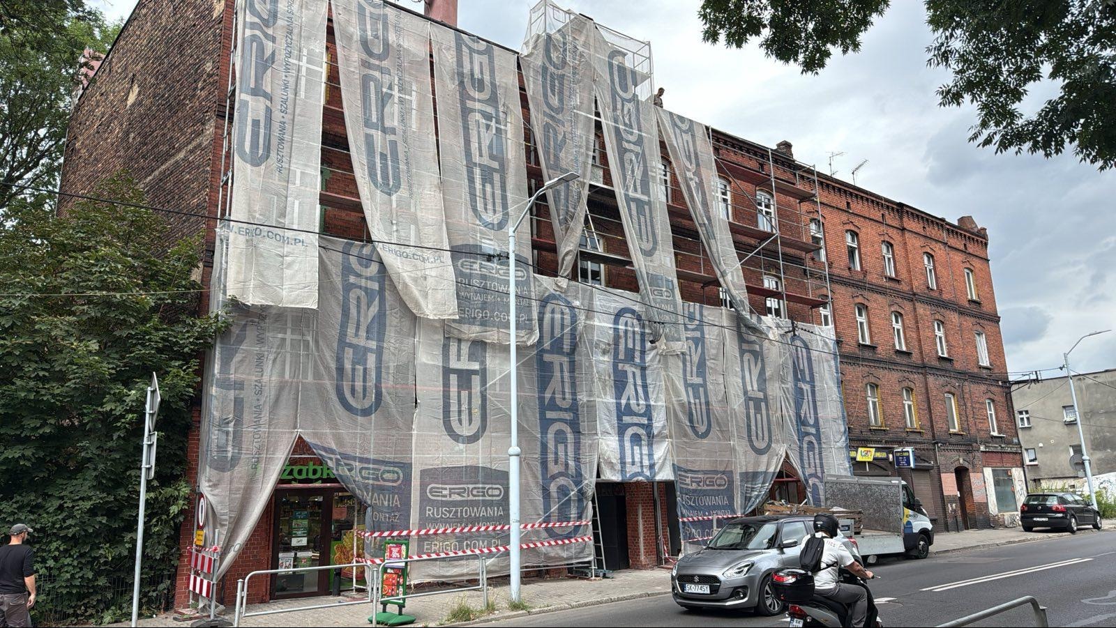 Budynek w rusztowaniach, osłonięty banerami z logo firmy. Widoczne fragmenty elewacji z czerwonej cegły. Na pierwszym planie ulica z przejeżdżającymi pojazdami.