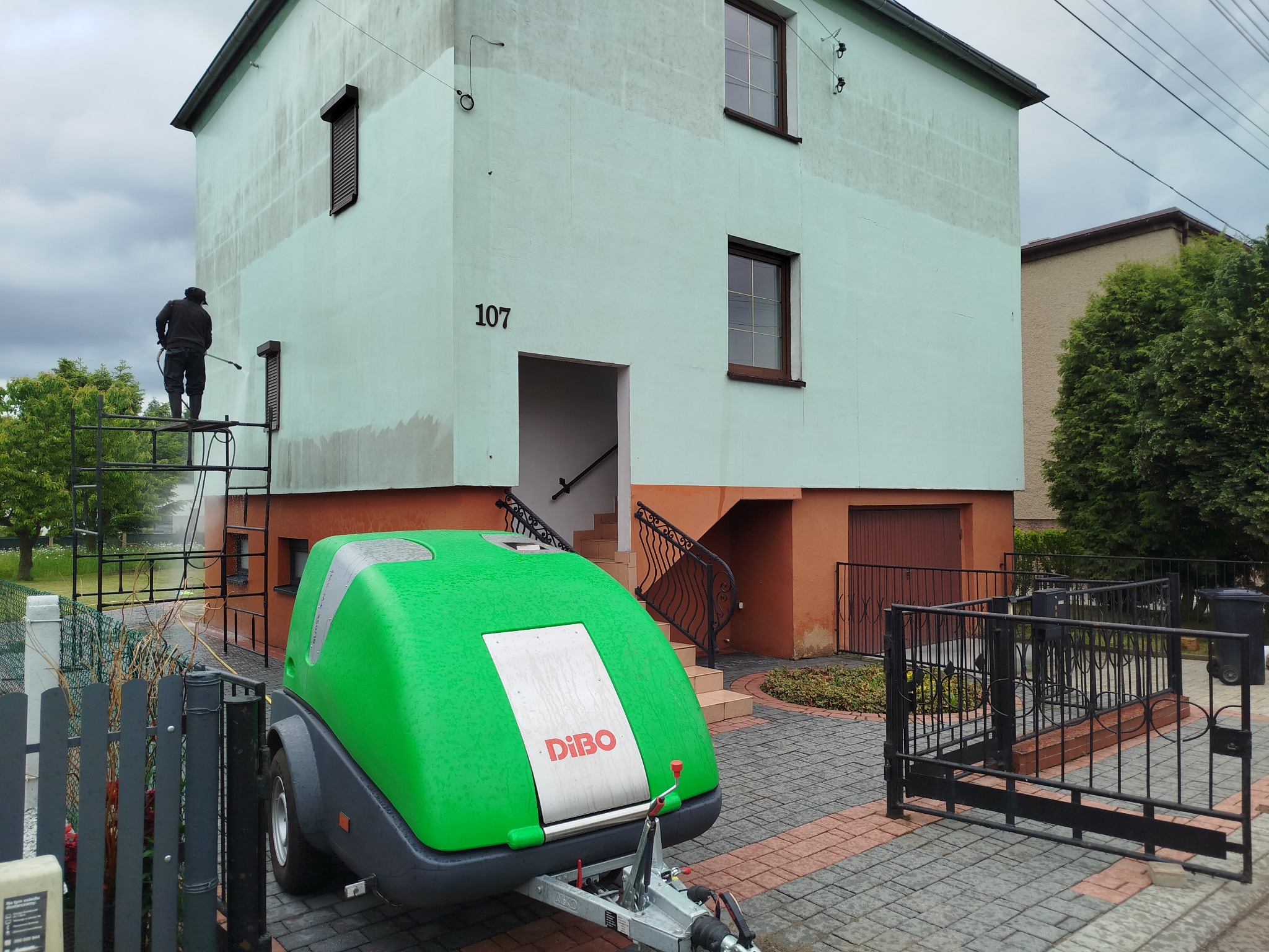 Pracownik na rusztowaniu czyści elewację domu z numerem 107 za pomocą myjki ciśnieniowej Dibo. Widoczny efekt czyszczenia - górna część elewacji jest czystsza.