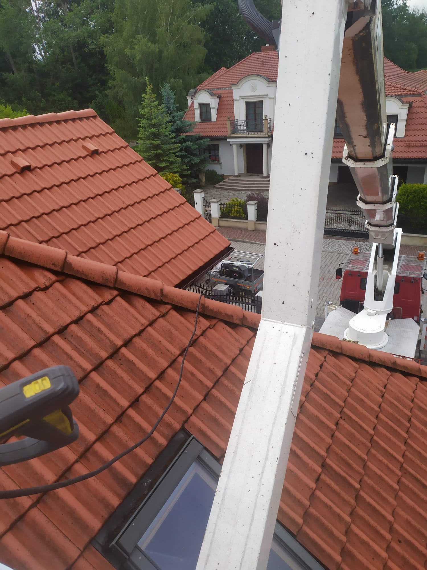 Czerwona dachówka widziana z góry, z fragmentem podnośnika koszowego i narzędziem do czyszczenia. W tle dom z ogrodem i drzewami.