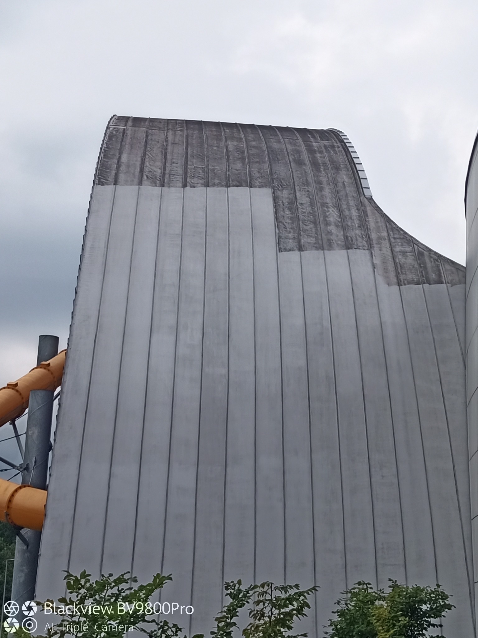 Częściowo umyta, wysoka elewacja budynku z zaokrąglonym dachem, widoczna z dołu. Obok żółta rura zjeżdżalni i zieleń. Szare niebo w tle.