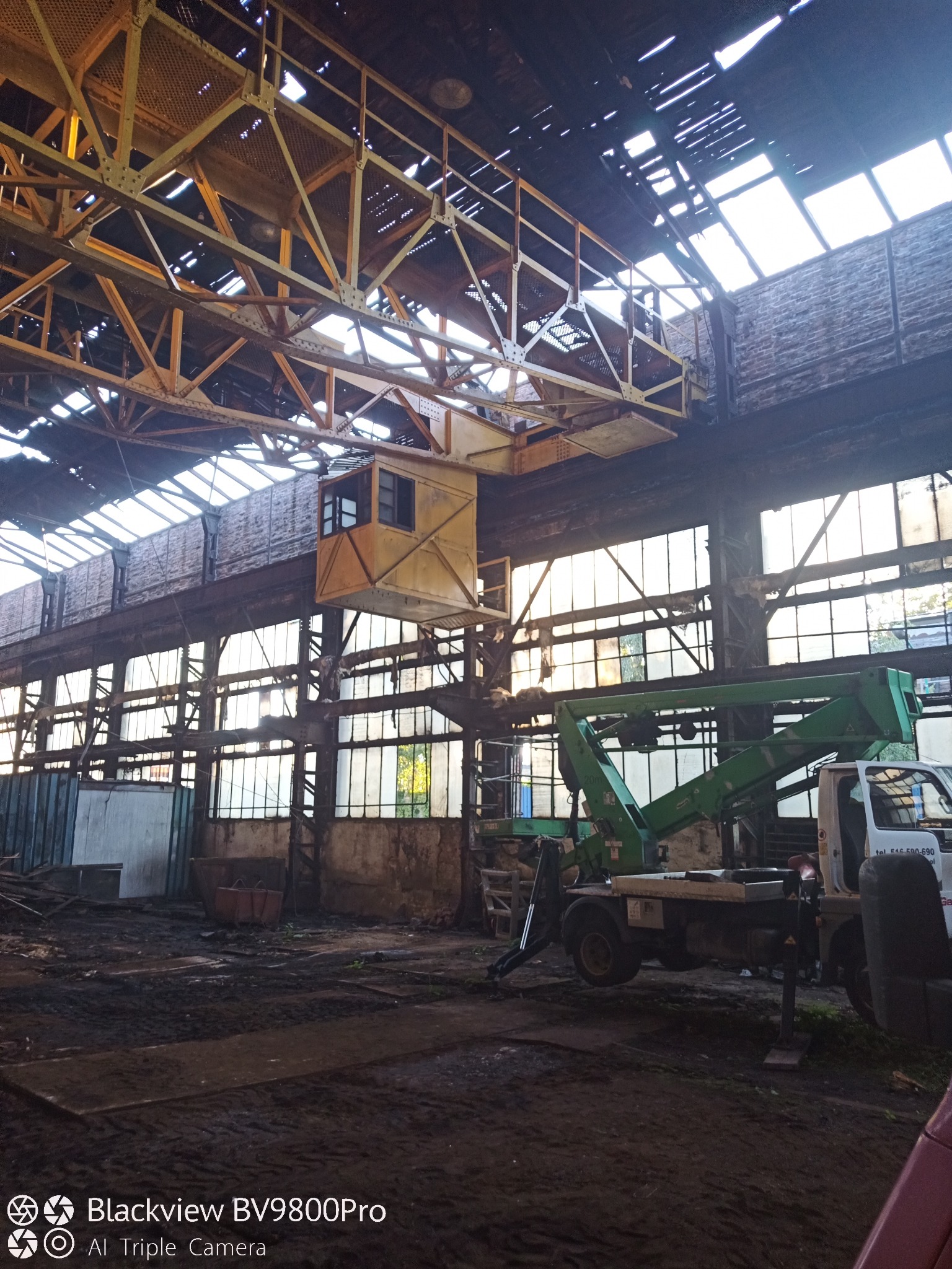 Wnętrze hali z suwnicą i podnośnikiem koszowym. Widoczne okna, konstrukcja dachu i podłoże. Oświetlenie naturalne, suwnica w kolorze żółtym, podnośnik zielony.