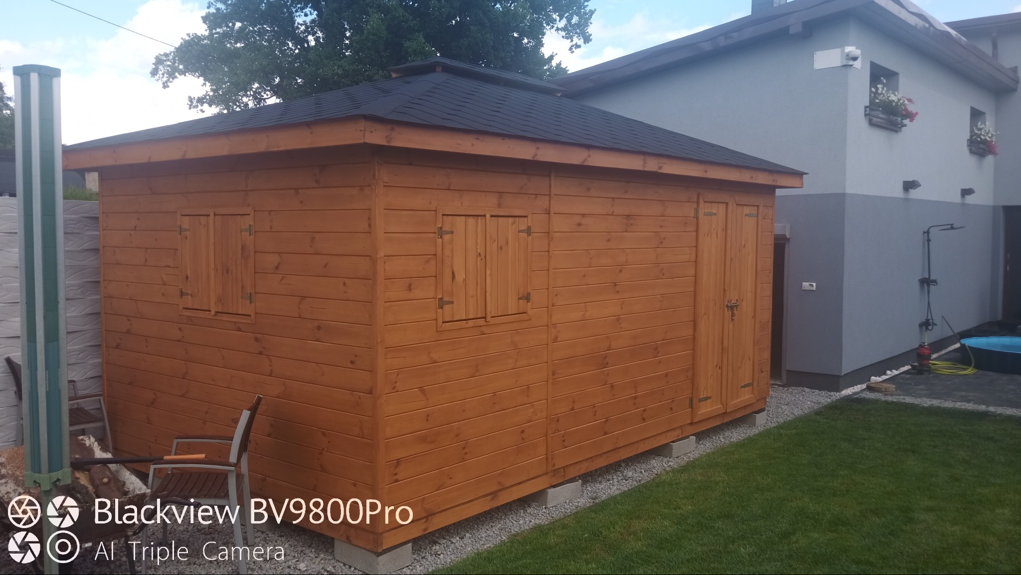 Drewniany domek narzędziowy z czarnym dachem, umieszczony na betonowych bloczkach, na tle trawnika i budynku mieszkalnego. Widoczne okiennice i drzwi.