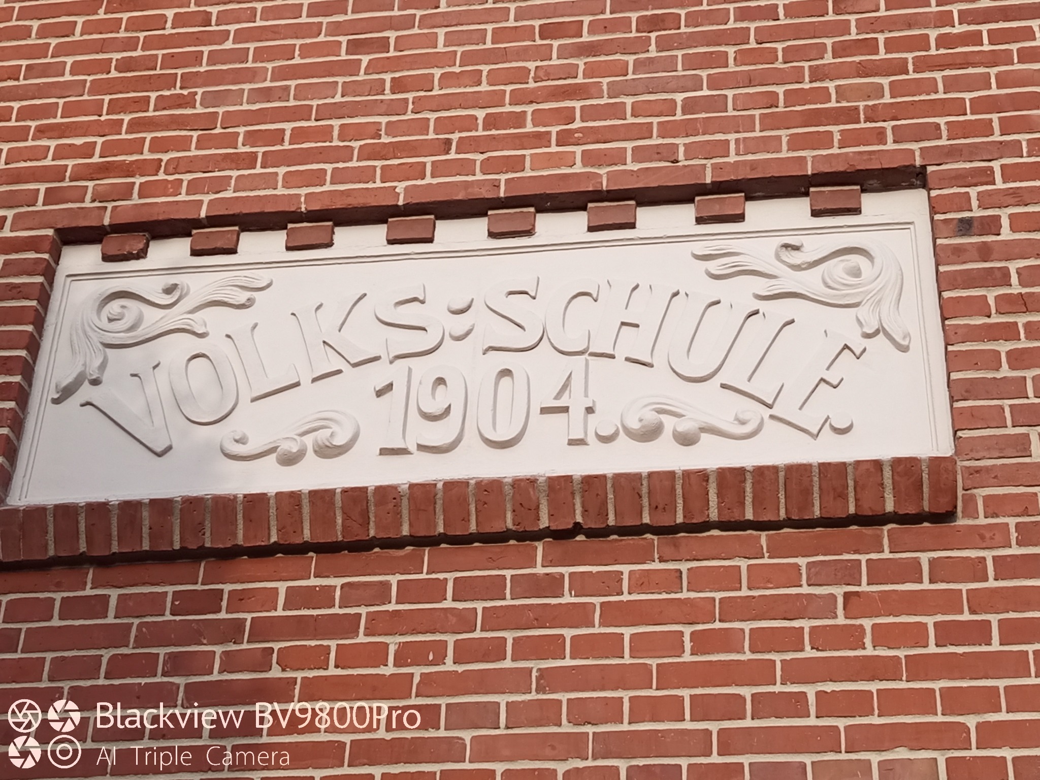 Tablica z napisem 'Volks:Schule 1904' na ceglanej ścianie budynku. Dekoracyjne elementy i białe litery na tle czerwonej cegły. Detal architektoniczny.