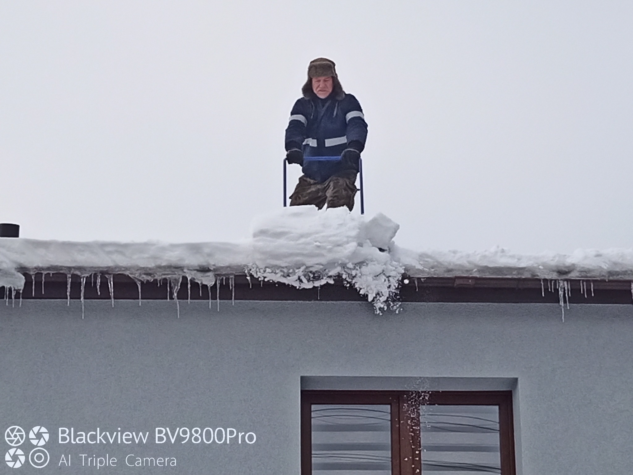 Mężczyzna w czapce usuwa śnieg z dachu domu za pomocą specjalnego narzędzia. Widoczne sople lodu i okno. Pochmurna pogoda.