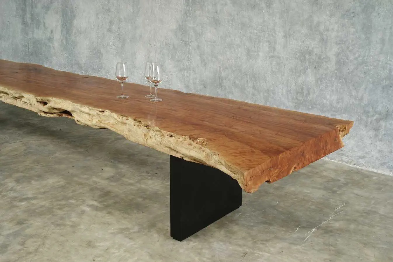 Unikatowy stół z litego drewna o nieregularnych krawędziach, wsparty na czarnej, minimalistycznej podstawie. Na blacie stoją trzy kieliszki do wina.