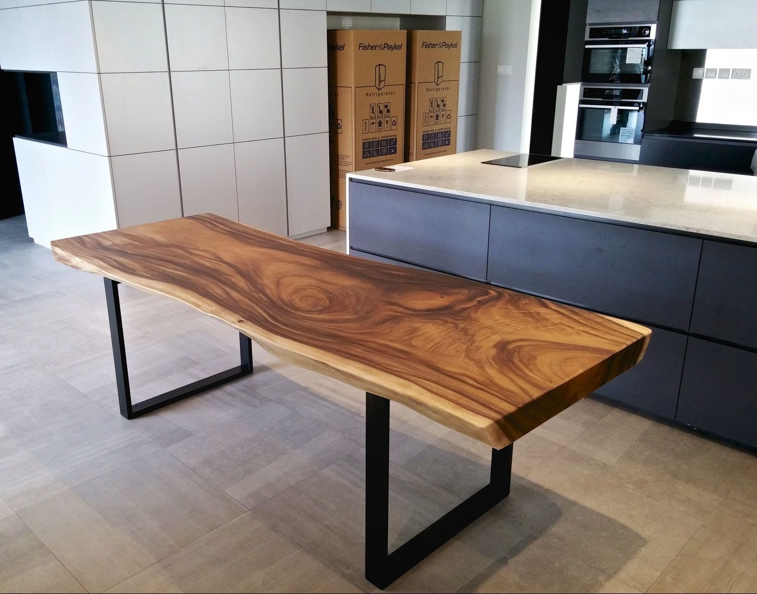 Stół z blatem z litego drewna o nieregularnych krawędziach, wsparty na czarnych, metalowych nogach w kształcie litery U. W tle nowoczesna zabudowa kuchenna.