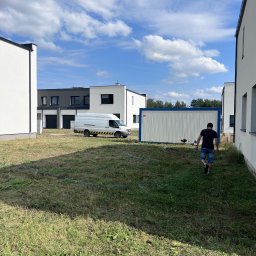 Twoja Przeprowadzka-Transport - Przygotowanie terenu: mężczyzna z kosiarką idzie w stronę kontenera budowlanego na zarośniętej działce między nowoczesnymi domami. Biały bus z żółtymi pasami w tle.