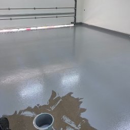 Twoja Przeprowadzka-Transport - Świeżo pomalowana, szara posadzka w garażu z otwartą bramą. Widoczne ślady czyszczenia i wiadro z wodą. Buty robocze w lewym dolnym rogu kadru.