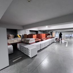 Twoja Przeprowadzka-Transport - Montaż mebli biurowych w garażu podziemnym. Rząd białych szafek z szufladami, elementy drewniane, pracownik w trakcie montażu. Oświetlenie sufitowe, betonowa podłoga.